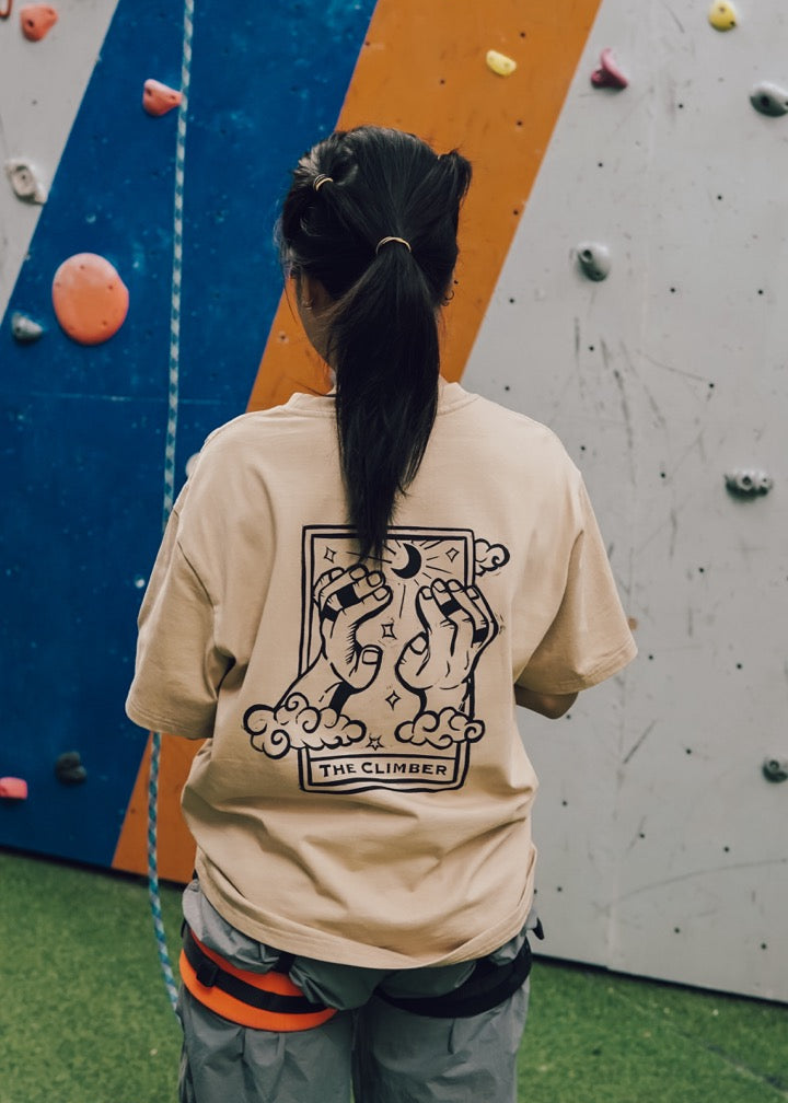 Person wearing a beige t-shirt with a graphic design, standing in front of a climbing wall.